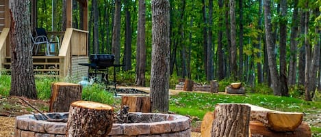 Una cabaña con terraza, un pozo de fuego y un bosque al fondo.