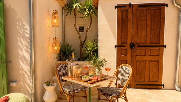Een terras buiten met een eettafel, rieten stoelen, een groene deur en een houten deur.