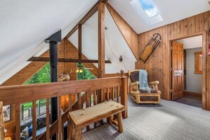 A loft bedroom with a sloped ceiling, wooden beams, a fireplace, a bench, a chair, and a view of greenery outside.