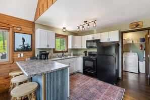 A kitchen with a central island, white cabinets, a black refrigerator, and a microwave. There are stools at the island and a window with a view of trees. - Elk Pointe Chalet- Hot Tub and Views! (Lead)