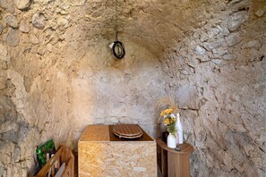 A rustic interior with a stone wall, a wooden table with a flat surface, and a small wooden stand holding a vase with flowers.