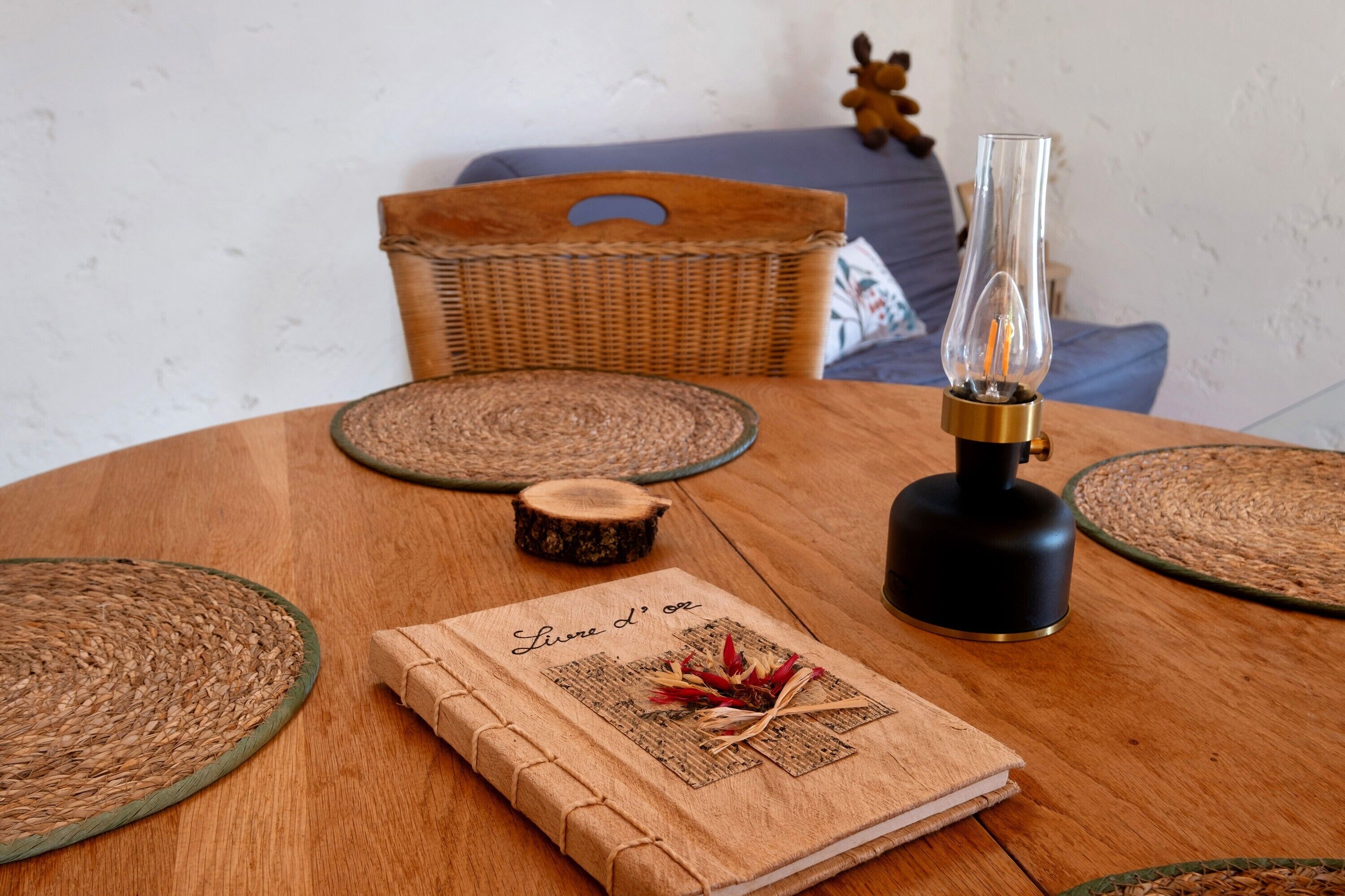 Une table ronde en bois, sur laquelle repose un set de table tissé, un petit sous-verre en bois, un porte-bougie en verre et un livre intitulé « Livre L’Ore ».