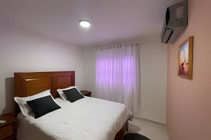 Ein Hotelzimmer mit einem Bett, Nachttischen, einer Deckenleuchte, einem Fenster mit Vorhängen und einer Wand-Klimaanlage.