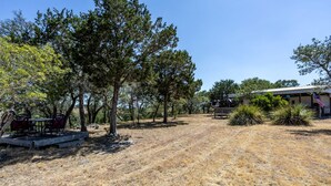 A dry grassy area with trees, a patio with chairs, and a building with an American flag. - Adventure Tent in Boerne, TX (Boerne)