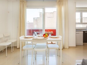 Un coin repas avec une table dressée pour deux personnes, des chaises blanches, une fenêtre avec des rideaux et une vue sur les bâtiments à l’extérieur.