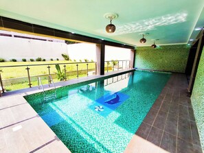 An indoor swimming pool with a clear view of the outside, a tiled floor, and a ceiling with recessed lighting. - L'Escale piscine et cinéma (Abidjan)
