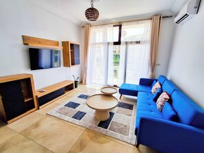 A modern living room with a blue sofa, a TV mounted on the wall, a wooden entertainment unit, and a large window with curtains. - L'Escale piscine et cinéma (Abidjan)