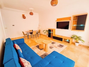 Een woonkamer met een blauwe bank, een eettafel met stoelen, een aan de muur bevestigde televisie en een potplant.