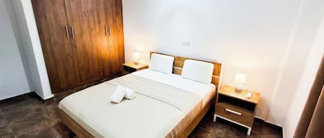 A bedroom with a bed, wooden wardrobe, bedside tables with lamps, and a wall-mounted light fixture.