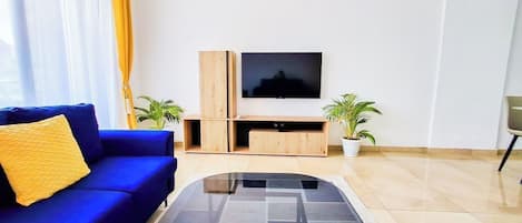 Un salon moderne avec un canapé bleu, une table basse en verre, un téléviseur posé sur un meuble en bois et deux plantes en pot.