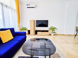 A modern living room with a blue sofa, a glass coffee table, a TV on a wooden stand, and two potted plants.
