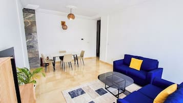 Een moderne woonkamer met een blauwe bank, een glazen salontafel en een eethoek met een tafel en stoelen.