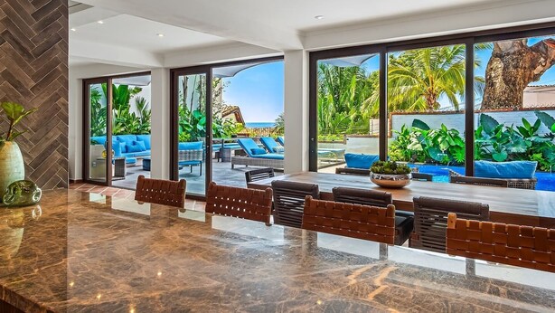 Een moderne hotellobby met een marmeren receptiebalie, grote ramen met uitzicht op een zwembad en tropische planten, en een zithoek met blauwe kussens.