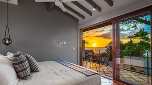 Een slaapkamer met een bed, kussens, een hanglamp en uitzicht op een ondergaande zon door een schuifdeur.