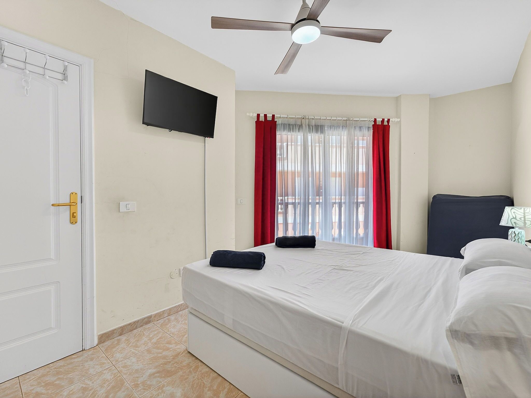 Hotelkamer met een bed, een flatscreen televisie, een plafondventilator en een raam met rode gordijnen.
