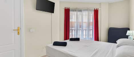 A hotel room with a bed, a flat-screen TV, a ceiling fan, and a window with red curtains.