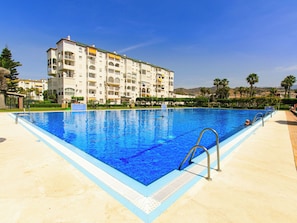 Ein Schwimmbecken mit glasklarer, blauer Wasseroberfläche, umgeben von einer gefliesten Liegefläche. Im Hintergrund ein mehrgeschossiges Gebäude mit Balkonen und einer flachen Dachterrasse.