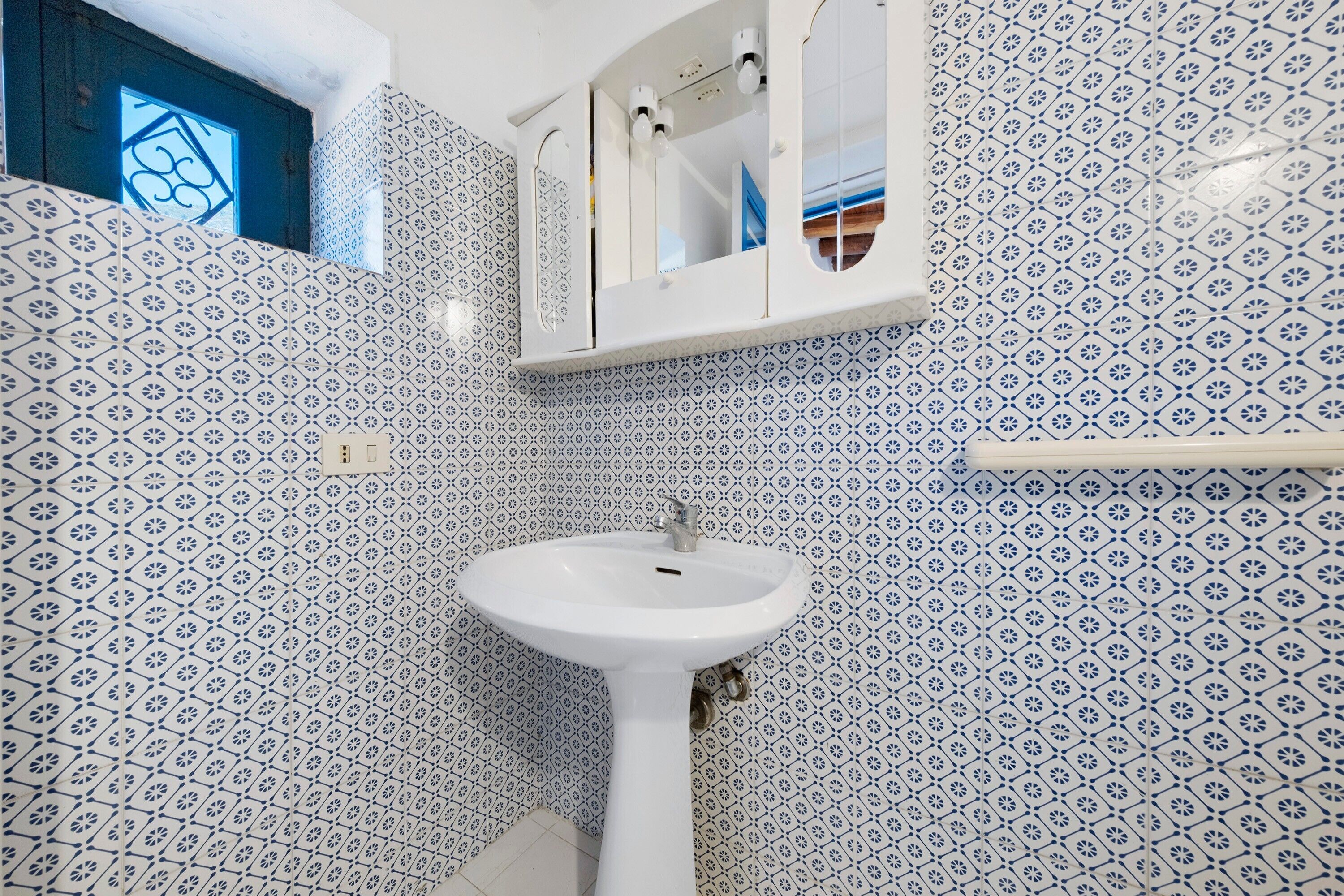 Ein Badezimmer mit einem weißen Waschbecken, blauen Fliesen mit Muster, einem Spiegel und einem Fenster mit geometrischem Design.
