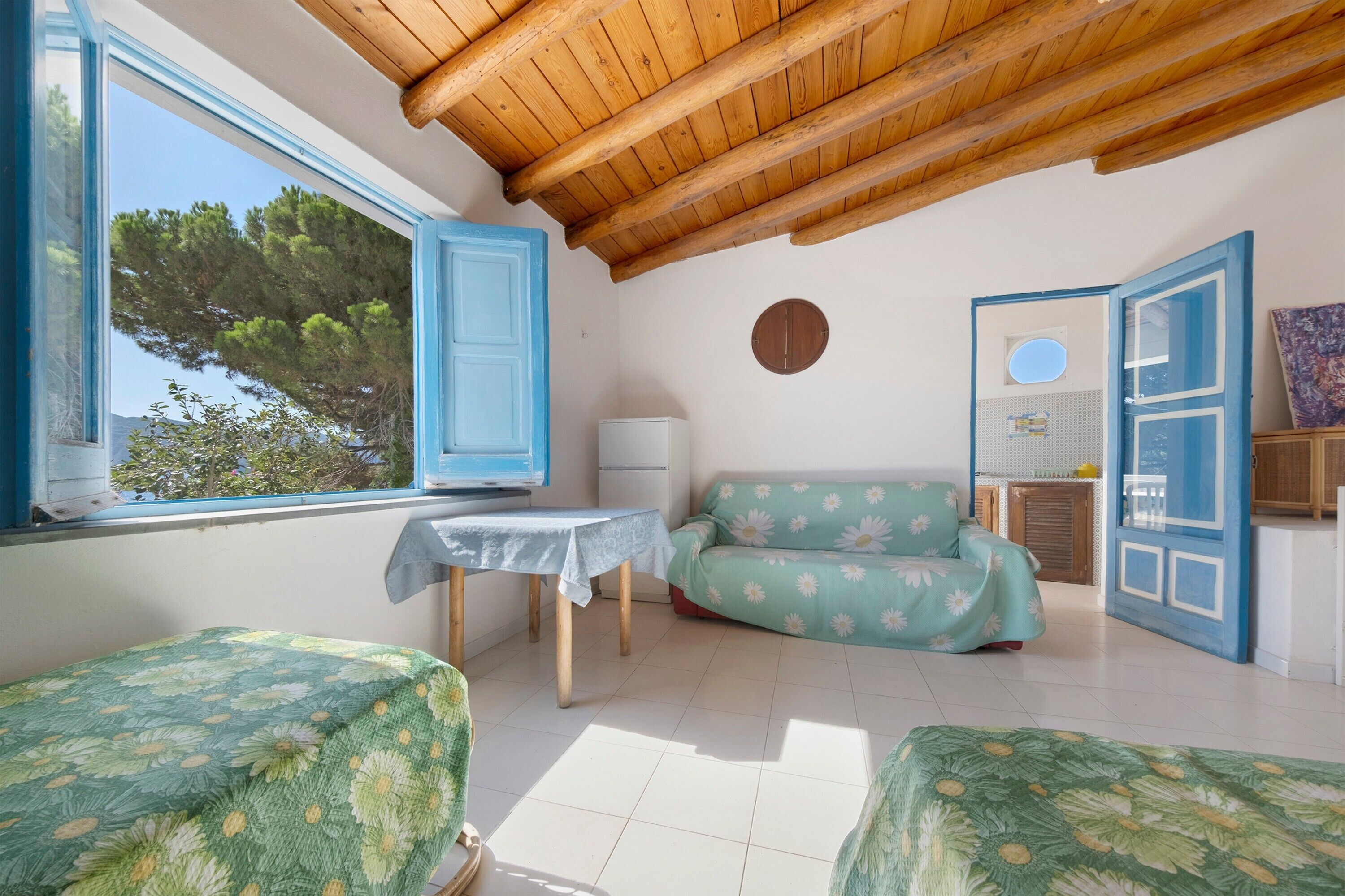 Una stanza con soffitto in legno, un divano con motivo floreale, un tavolo con tovaglia bianca e una finestra vista alberi.