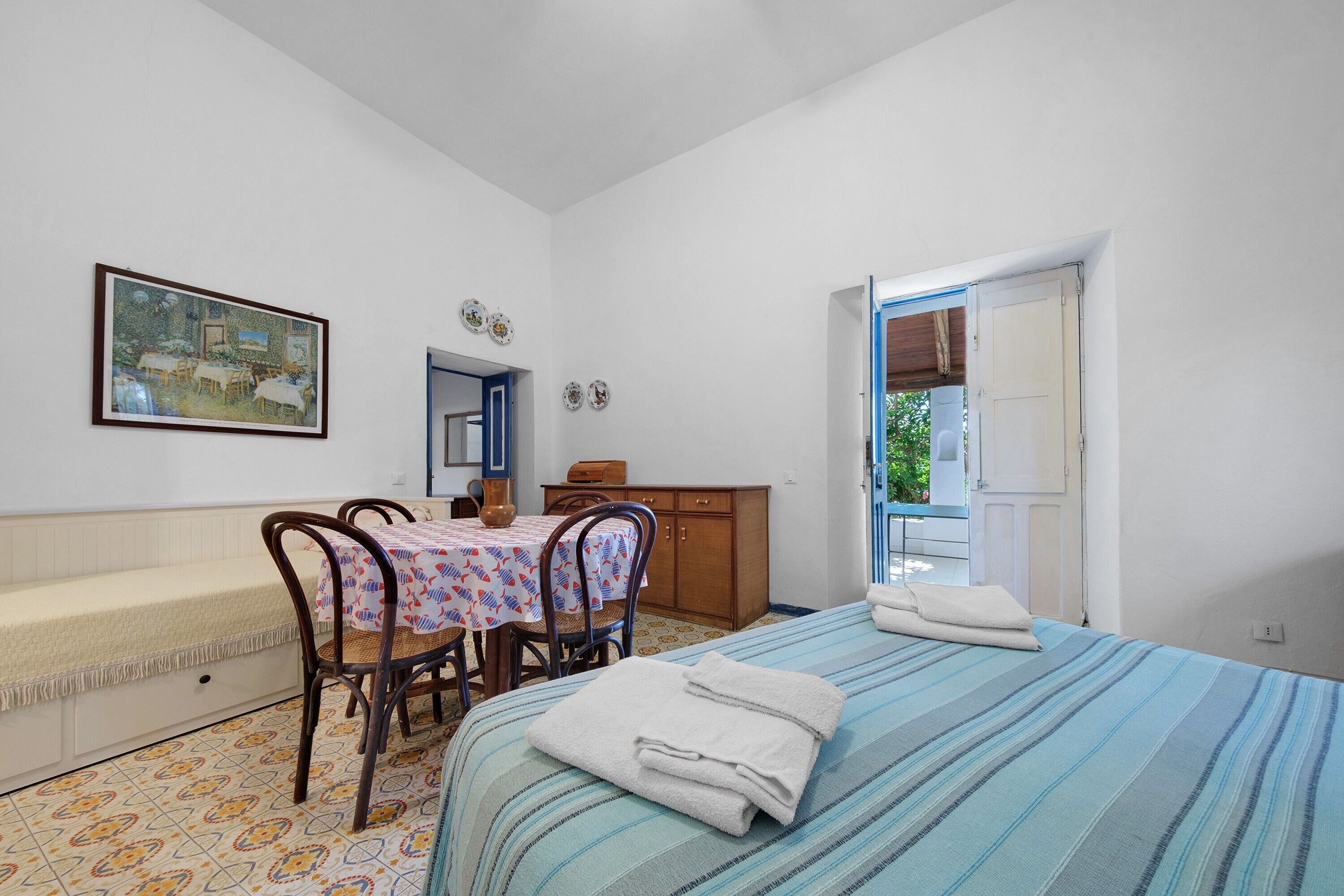 Una stanza con un letto, un tavolo con sedie, un quadro appeso al muro e una porta che dà sull'esterno.
