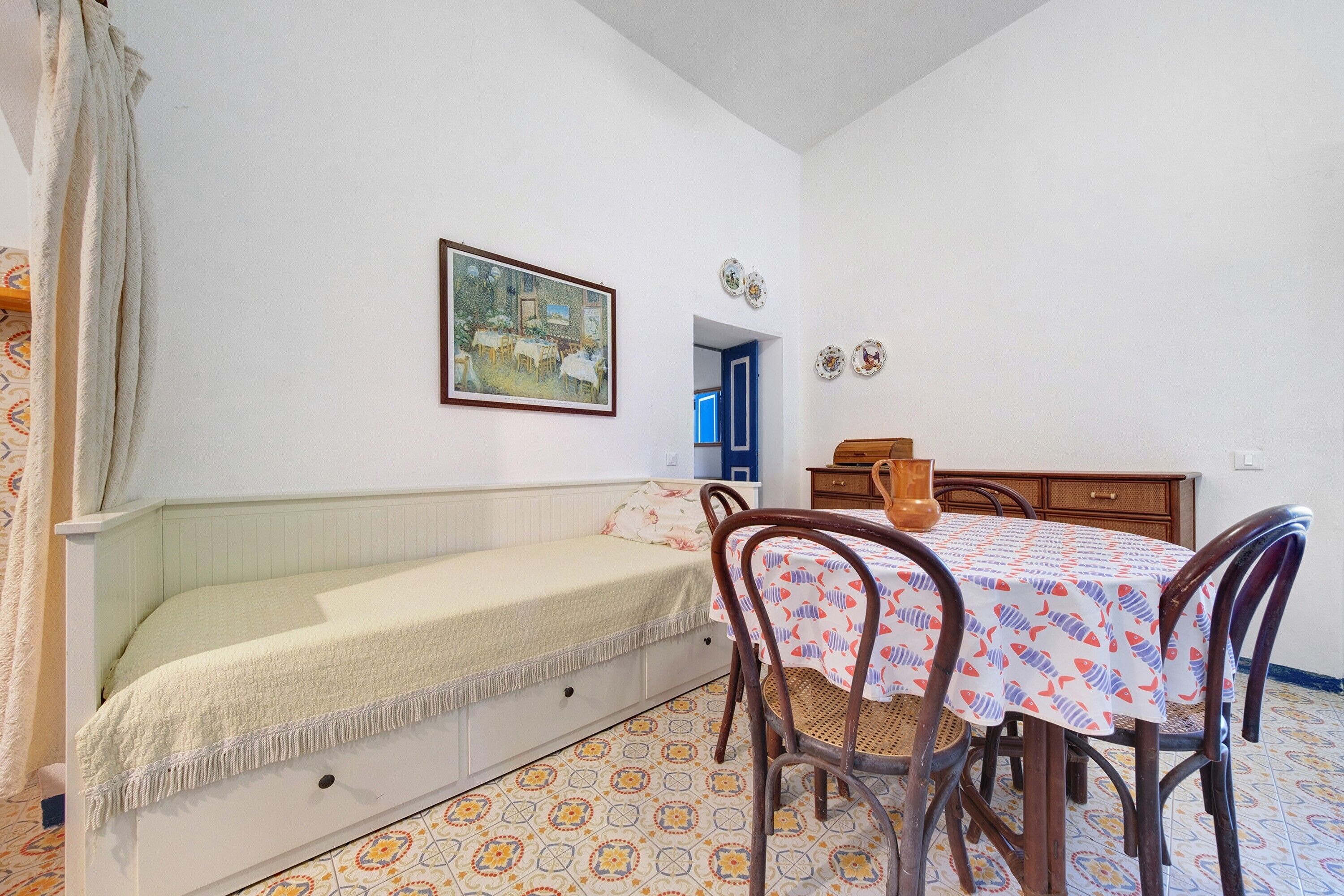 A room with a bed, a dining table with chairs, a painting on the wall, and a patterned floor.