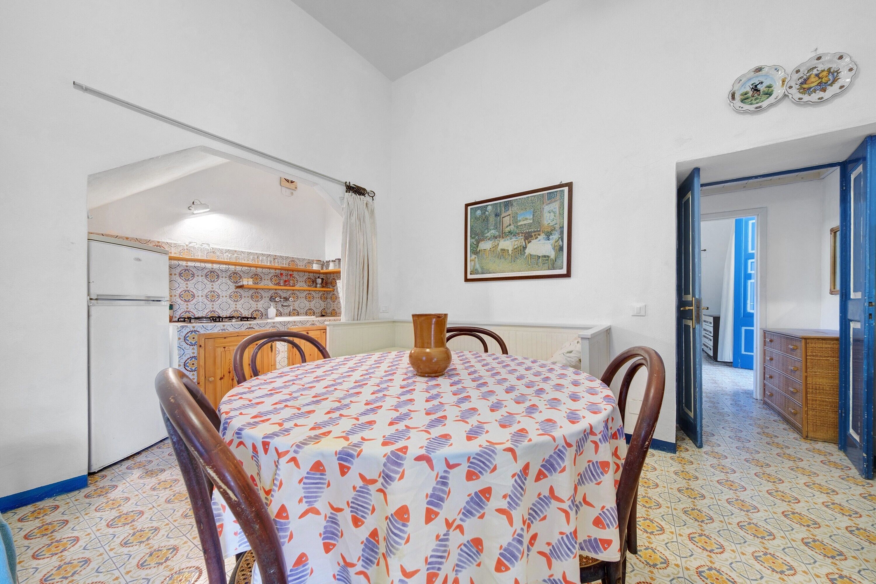 Un coin repas avec une table dressée pour quatre personnes, une cuisine en arrière-plan et une porte donnant sur une autre pièce.