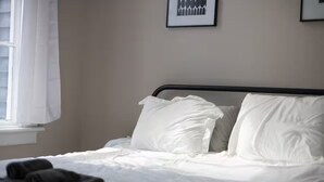 Ein Bett mit weißer Bettwäsche, ein Fenster mit weißen Vorhängen und ein gerahmtes Bild an der Wand.