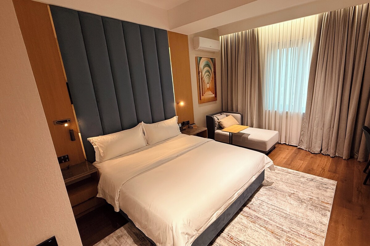 Quarto de hotel com uma cama grande, uma cama menor, uma escrivaninha e uma cadeira. Há um quadro na parede e uma janela com cortinas.