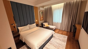 A hotel room with a large bed, a smaller bed, a desk, and a chair. There is a painting on the wall and a window with curtains. - PAZIRO HOTEL (Dushanbe)