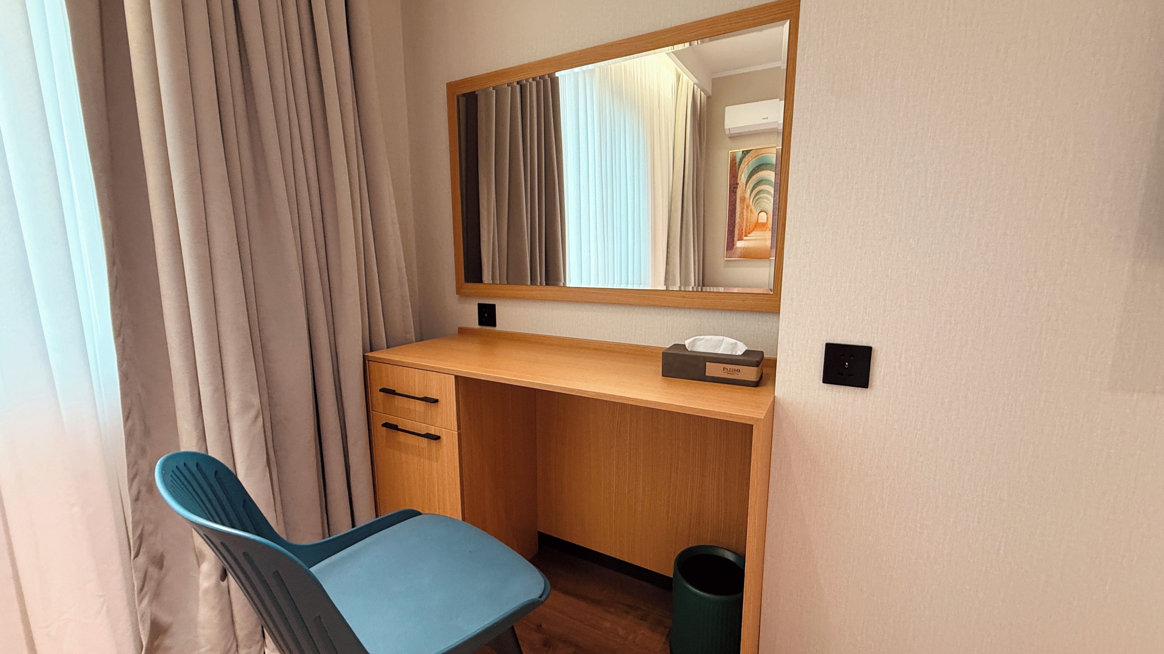 A compact room with a wooden desk, a chair, a mirror, and a trash bin.