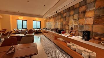 A hotel dining area with tables, chairs, a buffet counter, and a city view through large windows.