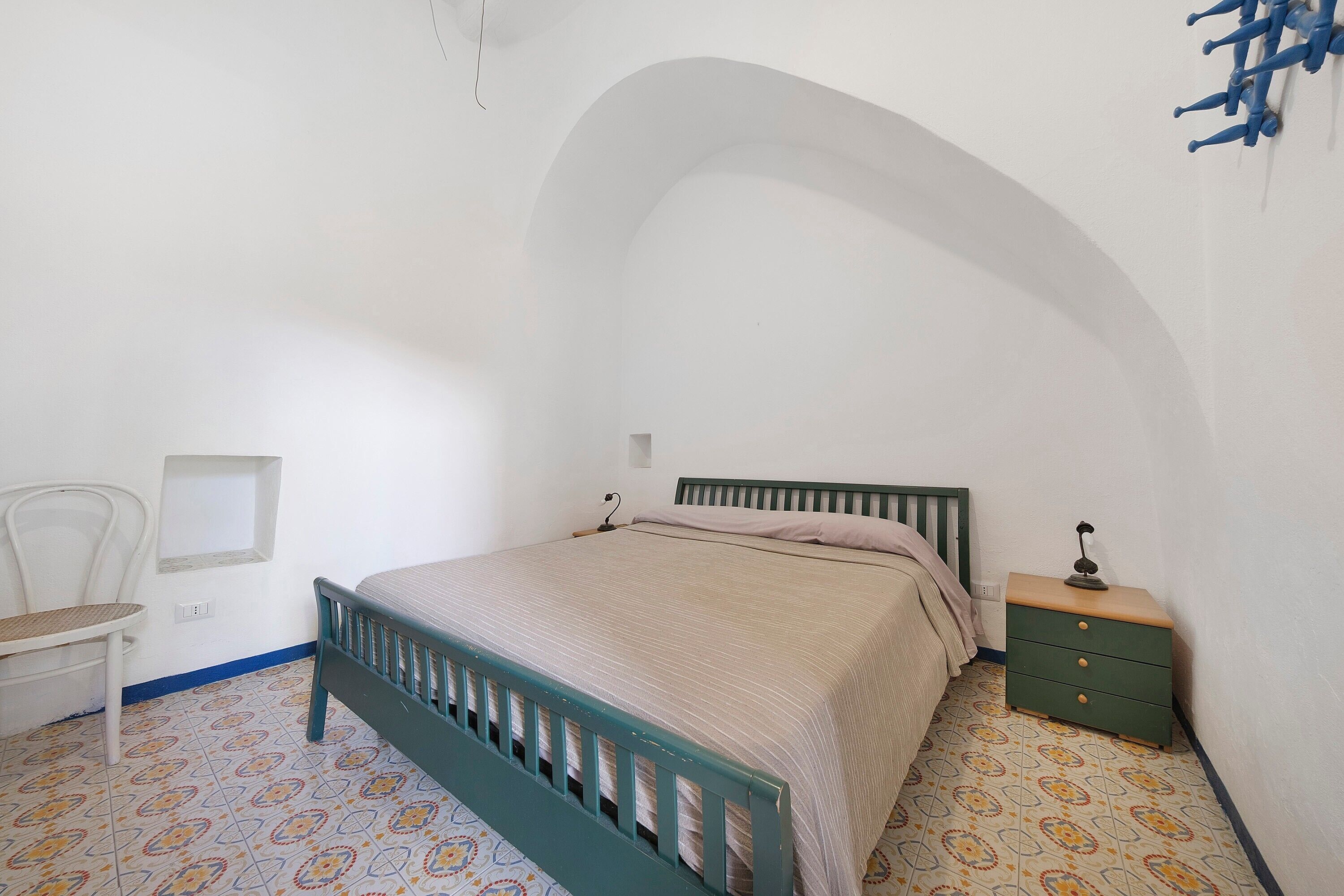 A bedroom with a single bed, a chair, a nightstand, and a wall-mounted lamp.