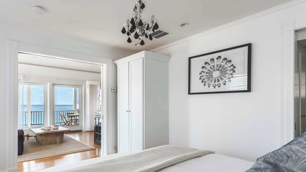 Un dormitorio con cama, candelabro, un cuadro enmarcado y vista al océano a través de amplias ventanas.