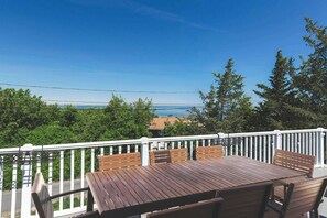 Eine Holzterrasse mit Tisch und Stühlen, einem weißen Geländer und Blick auf das Meer und Bäume.