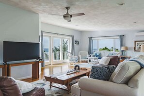 A spacious living room with a flat-screen TV, a ceiling fan, and a large window offering a view of the outdoors. - Stay at Wingaersheek (Gloucester)