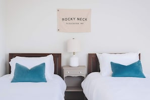 Une chambre d’hôtel avec deux lits, une table de chevet avec une lampe et une pancarte au-dessus du lit indiquant : « ROCKY NECK, Gloucester, Massachusetts ».