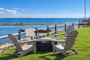Un espace aménagé au bord de l’eau, avec des fauteuils Adirondack et une table ronde, d’où l’on peut admirer un plan d’eau avec des bateaux et une jetée en bois.