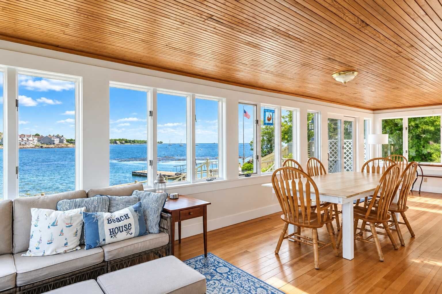 Un espacio amplio con una gran mesa de comedor, sillas de madera, un sofá con cojines estampados y amplias ventanas con vista al mar y a un muelle.