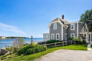 Une maison à deux étages, à l’extérieur gris et aux bordures blanches, située au bord d’un plan d’eau, avec une clôture en bois et de la verdure au premier plan.