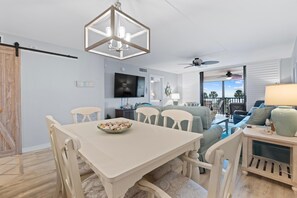 Ein Essbereich mit Tisch und Stühlen, einer Deckenleuchte, einem Fernseher und einer Couch im Hintergrund.