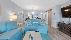 A living room with a blue sofa, a coffee table, and a television. - Caper Beach Club 313 (Fort Myers Beach)