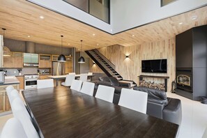 Eine moderne Küche mit Holzausstattung, ein Essbereich mit großem Tisch und weißen Stühlen, eine Treppe, ein Wohnzimmer mit schwarzem Ledersofa und einem Kamin.