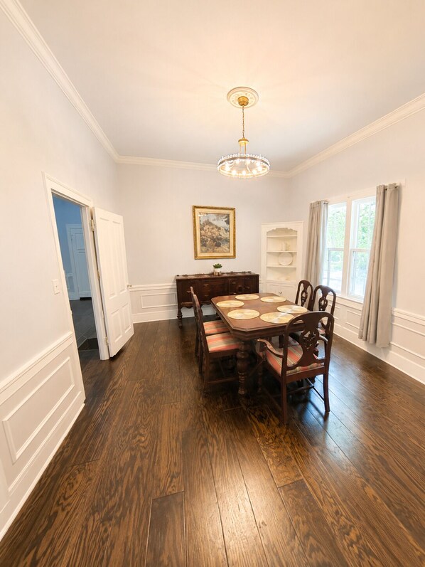 A dining room with a wooden table and chairs, a chandelier, a painting on the wall, and a window with curtains.