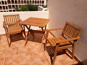 Ein Holztisch und zwei Stühle auf einer gefliesten Terrasse, im Hintergrund eine weiße Wand und ein Gebäude mit Bögen.
