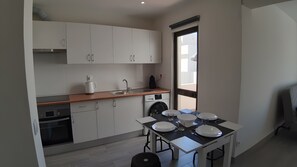 A compact kitchen with a sink, stove, and dishwasher, a dining table with plates, and a window providing natural light. - Dream Studio with Pool in Poente da Aldeia (Albufeira)
