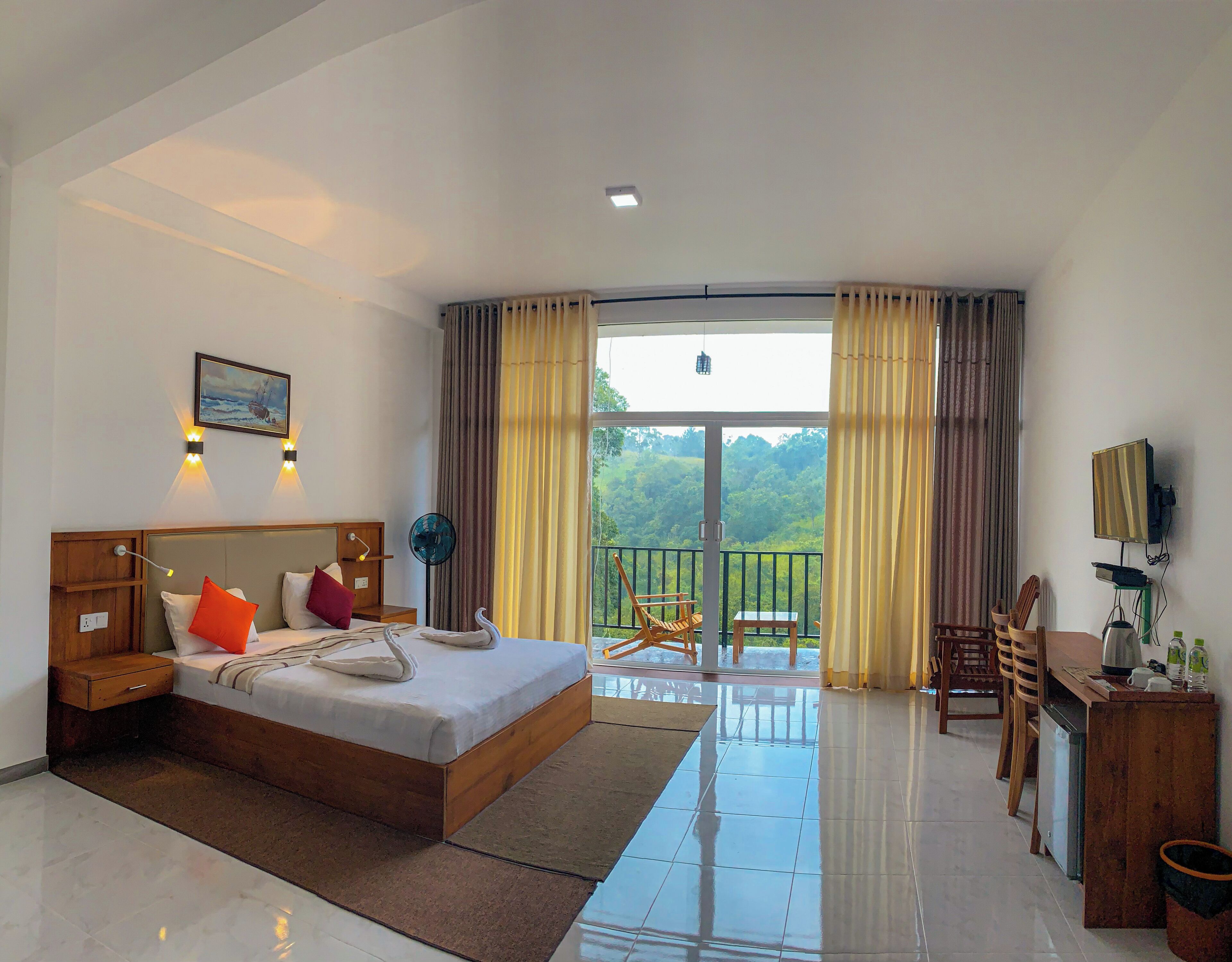 A hotel room with a bed, a desk, a chair, a fan, and a view of the outdoors.