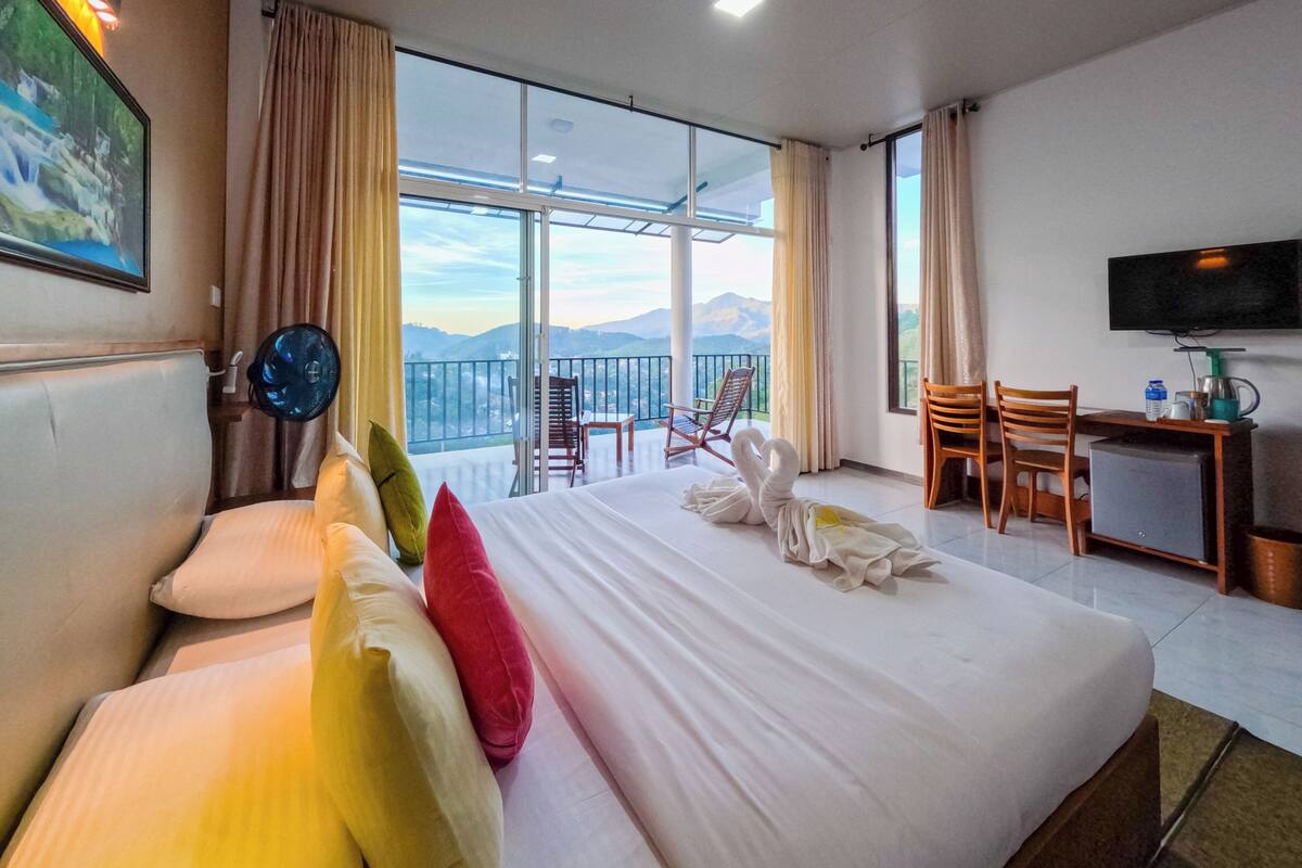Habitación de hotel con una cama grande, un escritorio, una silla, un televisor y un balcón con vistas a la montaña.