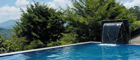 A swimming pool with a waterfall feature, surrounded by lush greenery and a clear sky.