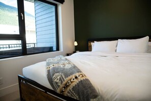 A bedroom with a bed, a window with a view of a building and mountains, a bedside table with a lamp, and a dark headboard. - Revy Rendezvous | Private Hot Tub & Mountain Views (Revelstoke)