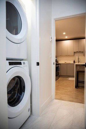 A laundry room with two front-loading washing machines and a kitchen with light wood flooring and white cabinets. - Revy Rendezvous | Private Hot Tub & Mountain Views (Revelstoke)